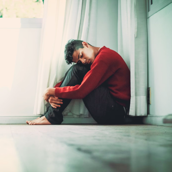 Image of a person sitting on the floor with their hands wrapped around their knees and their head resting on their knees to illustrate what stress and depression can look like